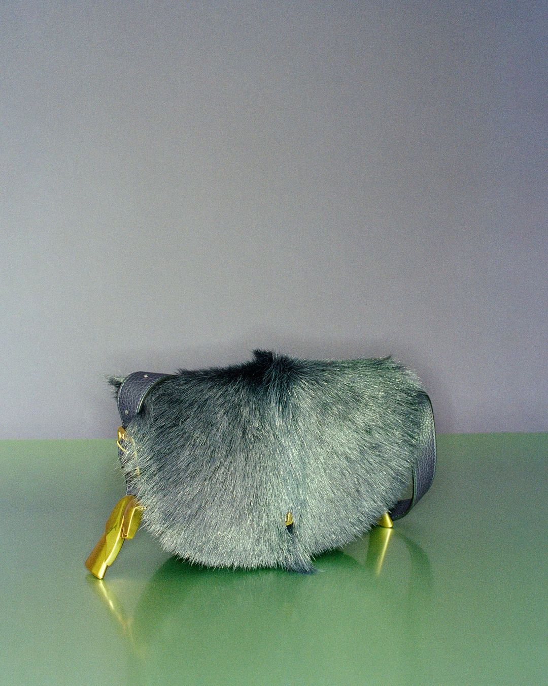 A whimsical blue faux fur clutch handbag with gold accents sits on a green surface against a two-tone blue-grey background.