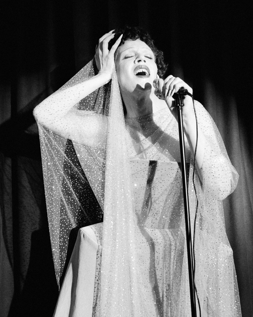 A woman in a sparkling dress and veil sings into a microphone, her hands raised in a dramatic pose against a dark background.