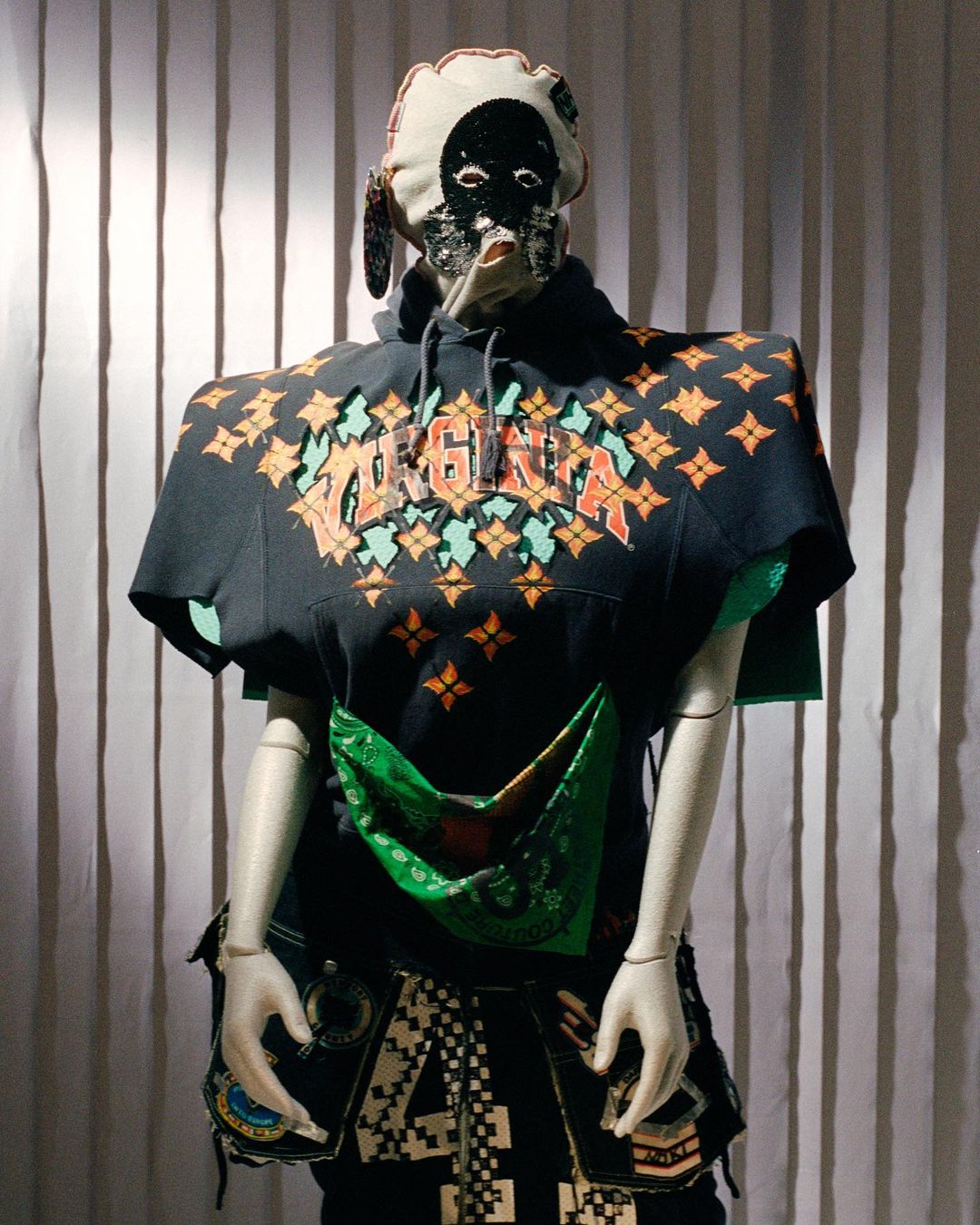 Mannequin in art exhibit with decorative black hoodie, matching mask, and colorful design elements in a bright, well-lit space.