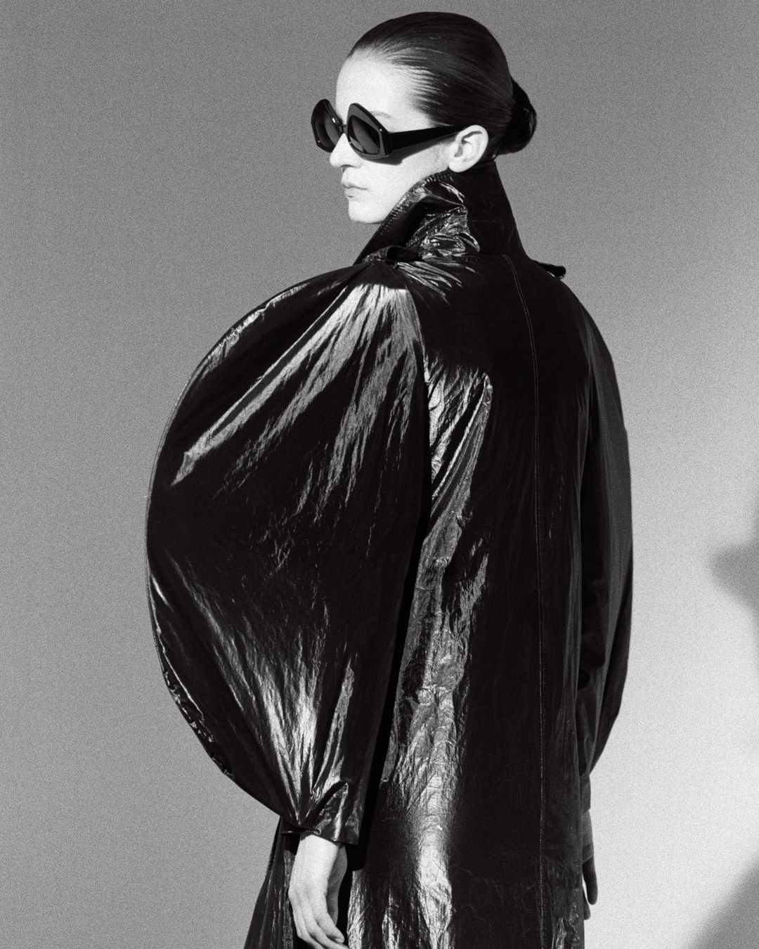 Black and white portrait of a woman wearing sunglasses and a shiny, puffy coat. Fashion shot in a photo studio with cool tone.