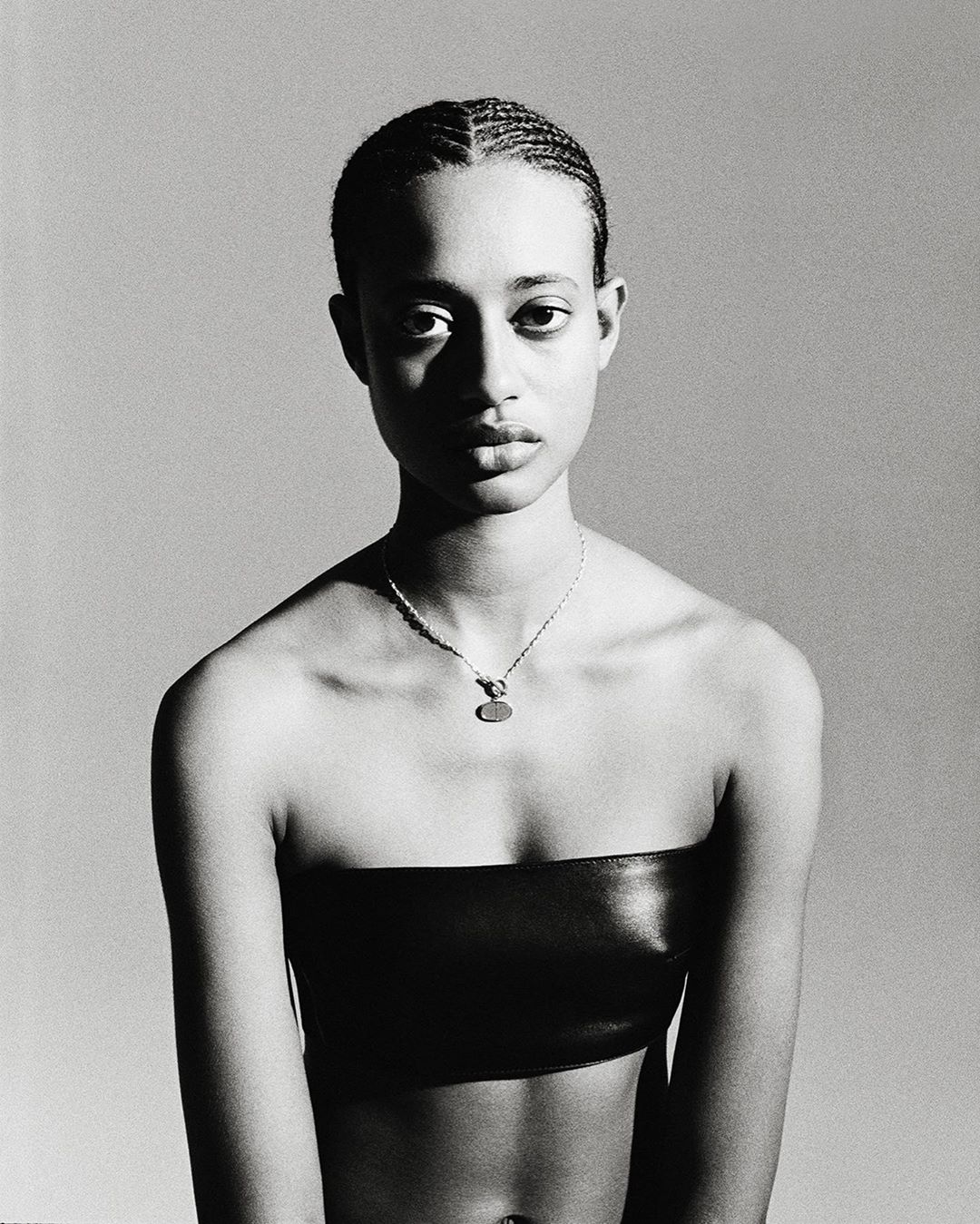 Portrait of a young adult woman with cornrow braids wearing a black bandeau top and pendant necklace against a grey backdrop.