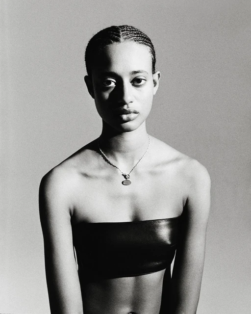 Portrait of a young adult woman with cornrow braids wearing a black bandeau top and pendant necklace against a grey backdrop.