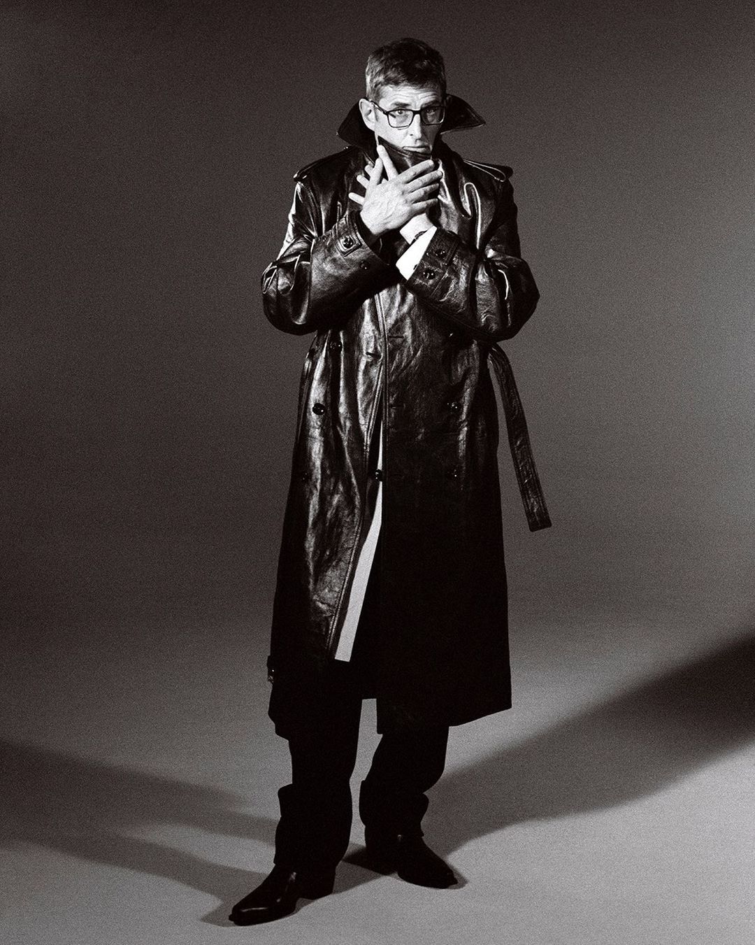 Man wearing a long dark leather trench coat and glasses, holding hands up to his face against a grey studio backdrop.