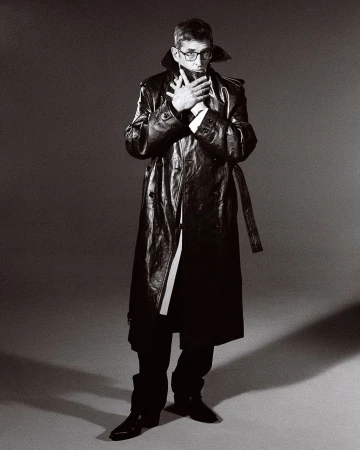 Man wearing a long dark leather trench coat and glasses, holding hands up to his face against a grey studio backdrop.