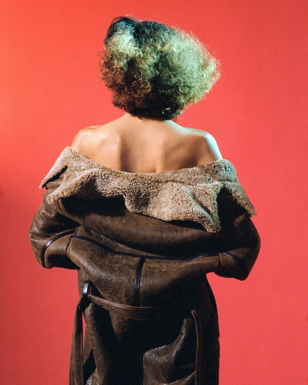 Back view of adult female with curly hair and brown shearling coat against a red backdrop in a studio environment with diffused lighting.