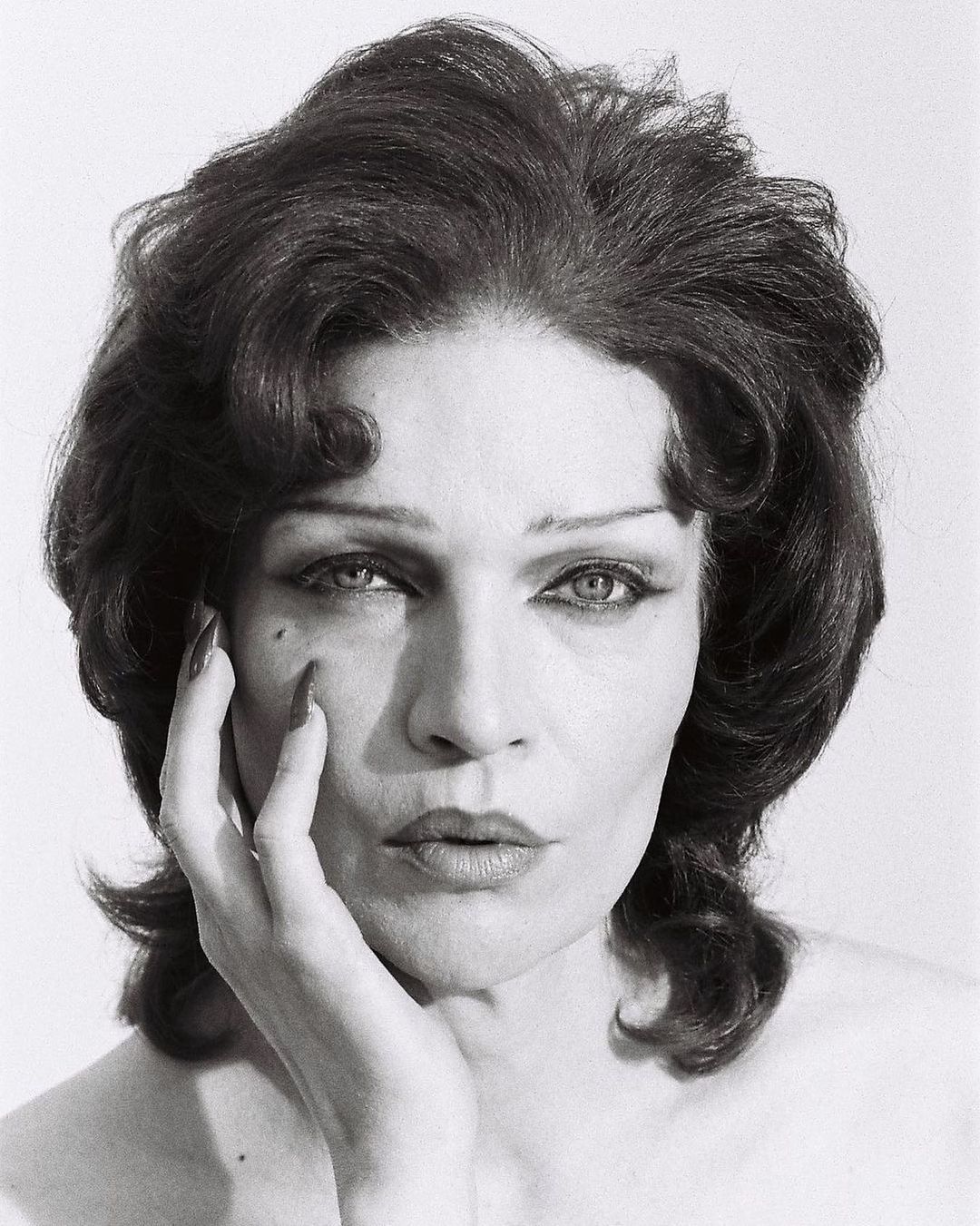 Monochrome portrait of an adult woman with dark curly hair, dramatic eye makeup, hand on her face, and a plain studio background.
