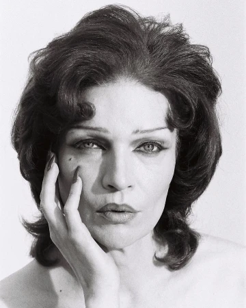 Monochrome portrait of an adult woman with dark curly hair, dramatic eye makeup, hand on her face, and a plain studio background.