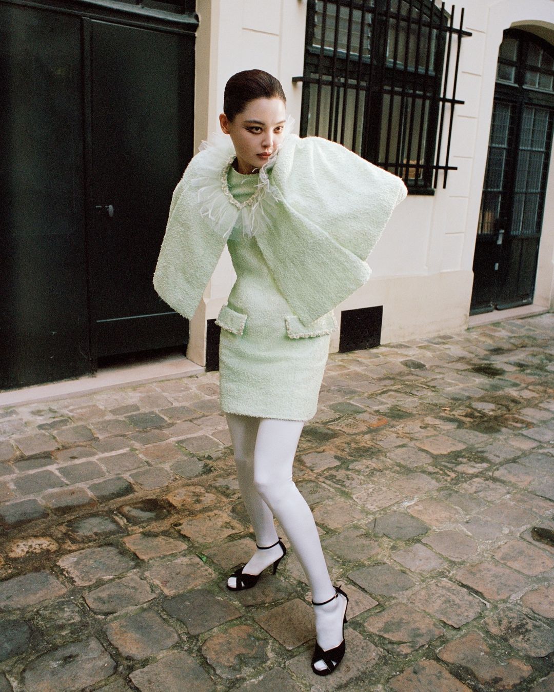 A young woman models a mint-green tweed dress with a feather collar and cropped jacket, paired with white tights and black heels, in an outdoor setting.