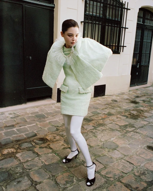 A young woman models a mint-green tweed dress with a feather collar and cropped jacket, paired with white tights and black heels, in an outdoor setting.
