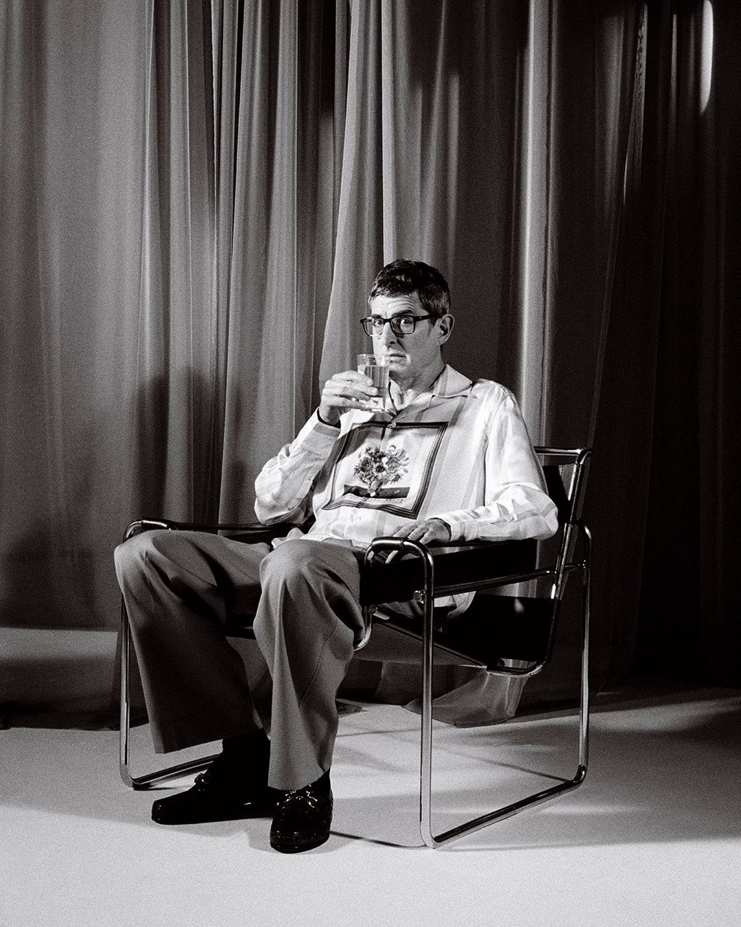 Monochrome portrait of a man with glasses seated in a modern chair, holding a glass, with a curtain backdrop in a studio setting.