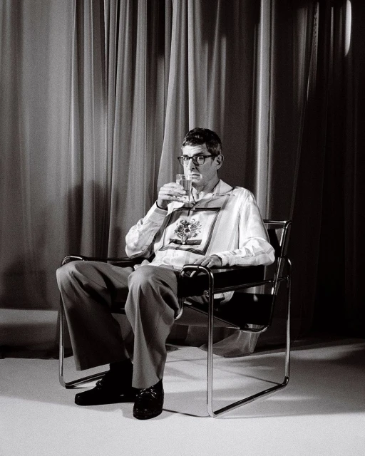 Monochrome portrait of a man with glasses seated in a modern chair, holding a glass, with a curtain backdrop in a studio setting.
