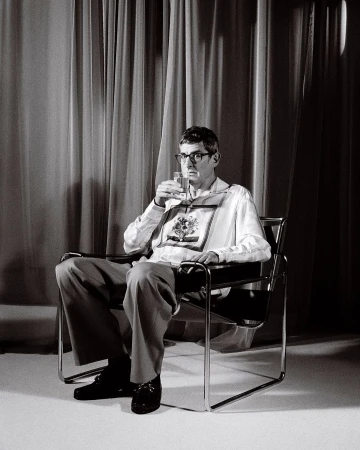 Monochrome portrait of a man with glasses seated in a modern chair, holding a glass, with a curtain backdrop in a studio setting.