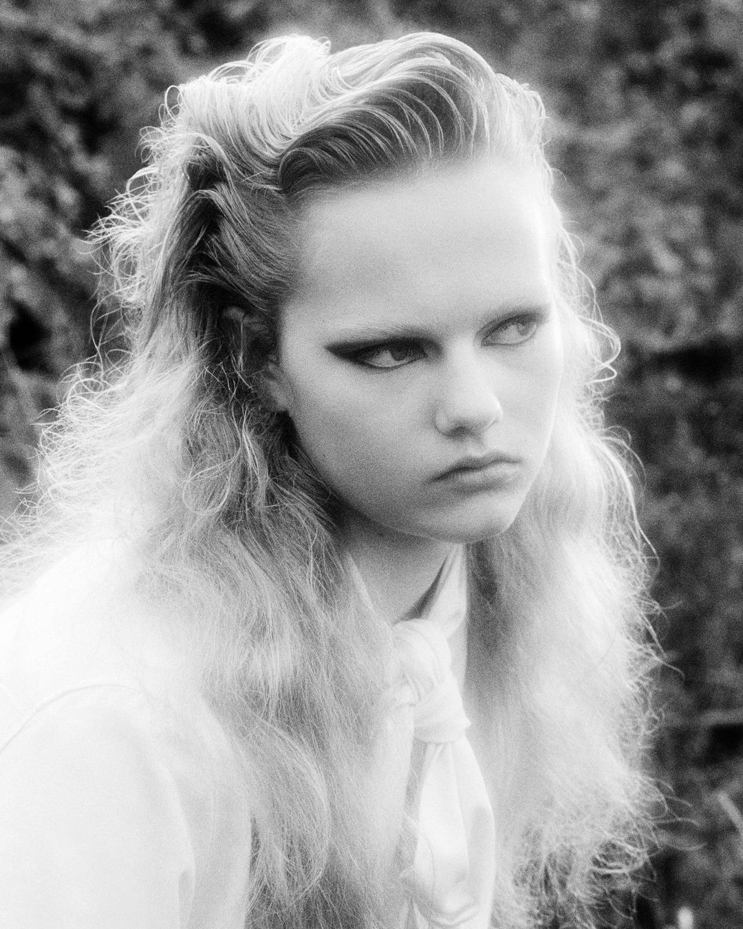 Monochrome portrait of an adult female with long blonde wavy hair, wearing a light-colored blouse, staring to the side against an outdoor background.