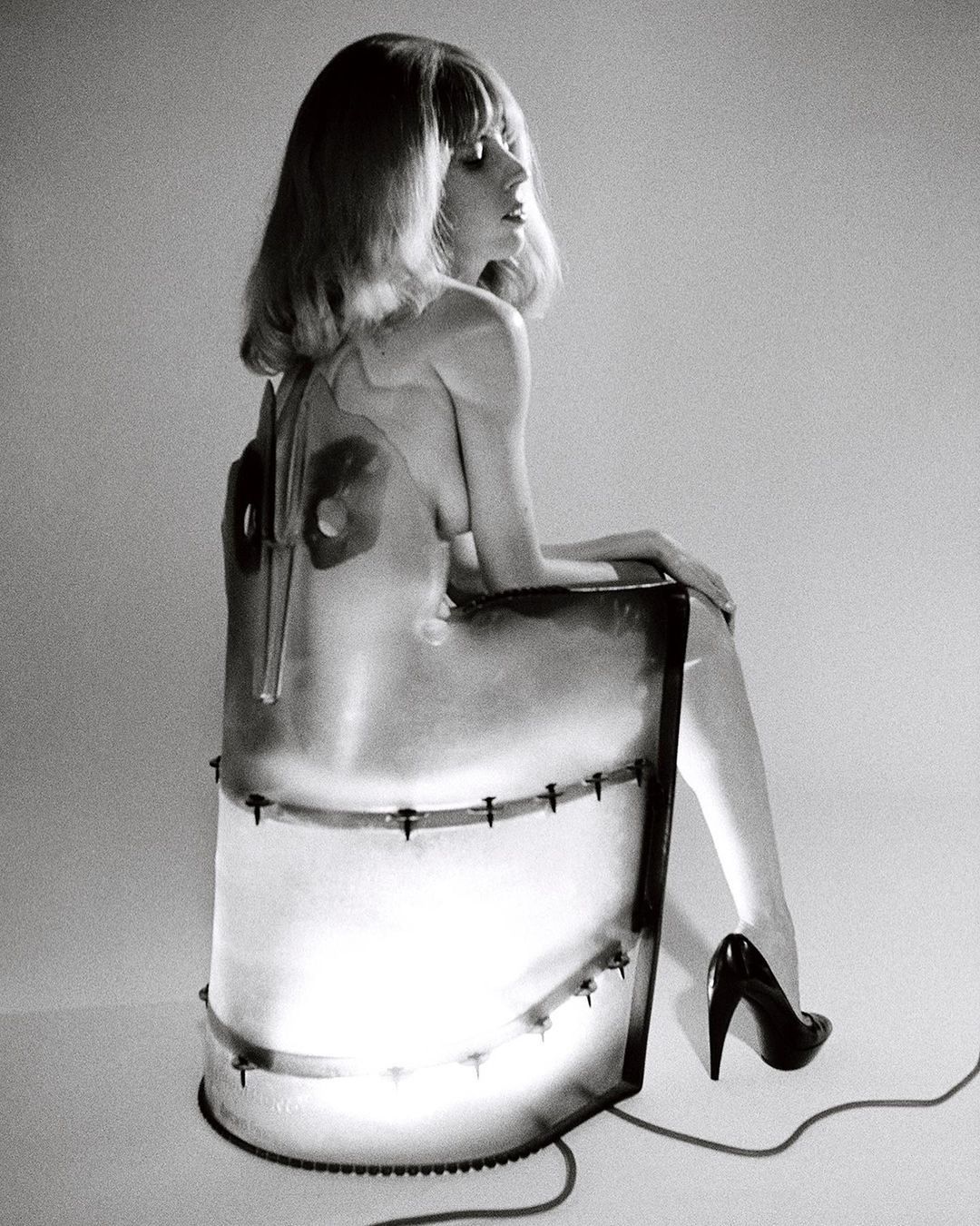 Black and white studio shot of a blond woman sitting on a chair, wearing high heels and a transparent dress with metal details.