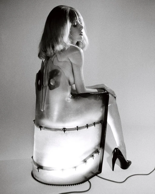Black and white studio shot of a blond woman sitting on a chair, wearing high heels and a transparent dress with metal details.