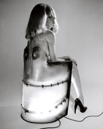 Black and white studio shot of a blond woman sitting on a chair, wearing high heels and a transparent dress with metal details.
