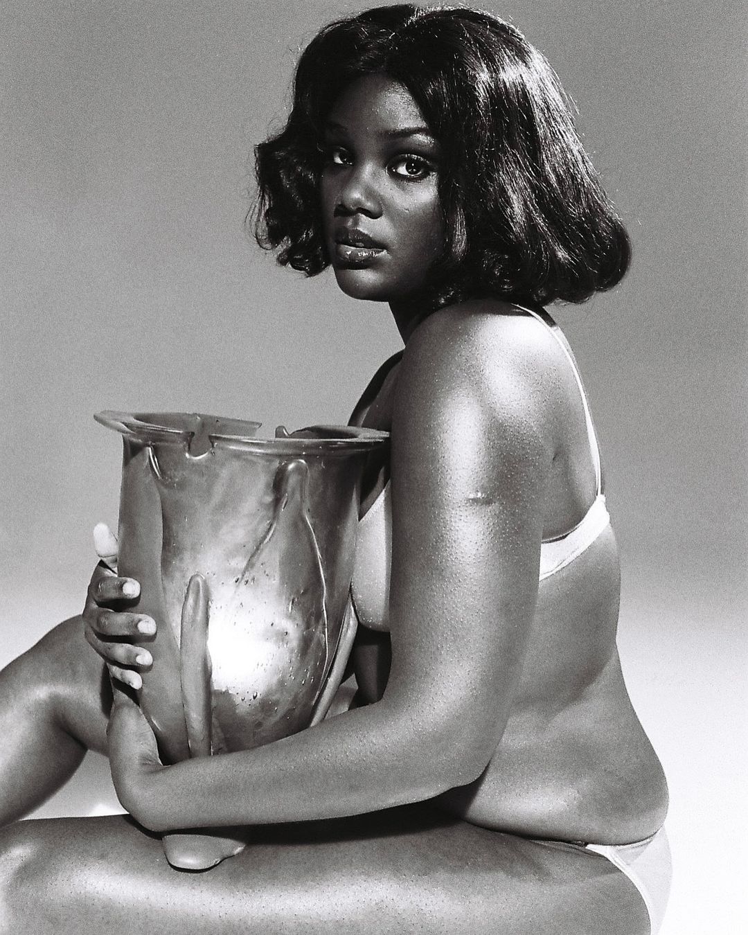 Black and white portrait of a young Black woman with shoulder-length hair sitting and holding a decorative metal vase in a photo studio.