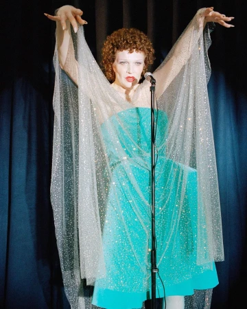 Red-haired woman in turquoise dress with sheer sleeves singing on stage in spotlight with microphone in front of dark blue curtains.