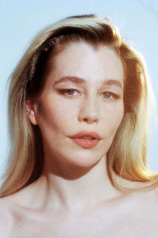 Portrait of a blonde woman with fair skin and light makeup against a light blue backdrop in soft lighting with a close-up camera angle.