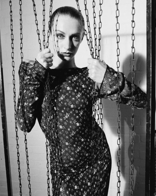 Monochrome portrait of a woman in a floral dress, posing with metal chains in front of a white brick wall in a photo studio.