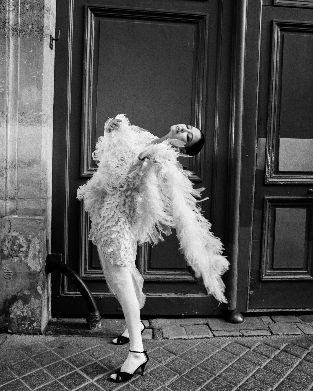 Fashionable woman poses in front of a weathered wooden door wearing an elegant feather dress and black heels in a black and white shot.