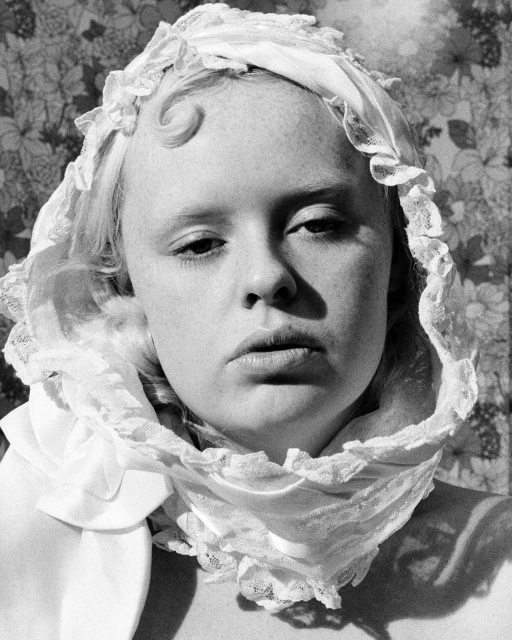 Black and white portrait of a fair-skinned adult female wearing a bonnet with lace trim, set against a floral patterned backdrop.