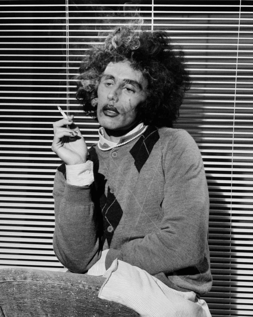 Monochrome portrait of an adult with afro hairstyle, wearing a cardigan, and smoking a cigarette in front of venetian blinds.