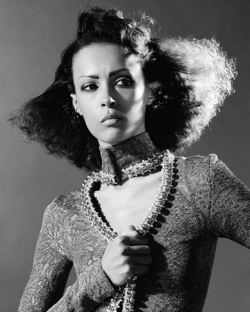 Monochrome portrait of a woman with textured dark hair wearing a high-collared patterned dress and holding a beaded necklace, shot by Camille Vivier