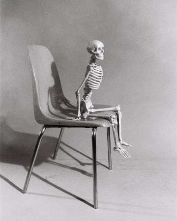 Monochrome photo of a human skeleton seated on a light-colored chair with metal legs, casting a shadow against a plain backdrop.
