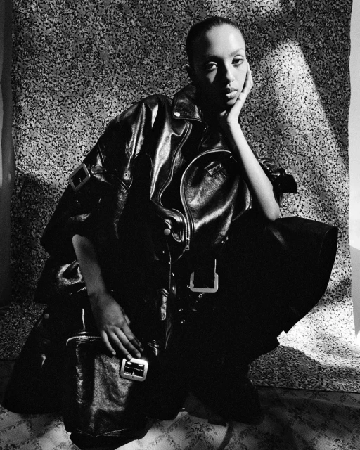 Monochrome portrait of an adult female model in a leather jacket, crouching against a patterned backdrop with a matching leather handbag.