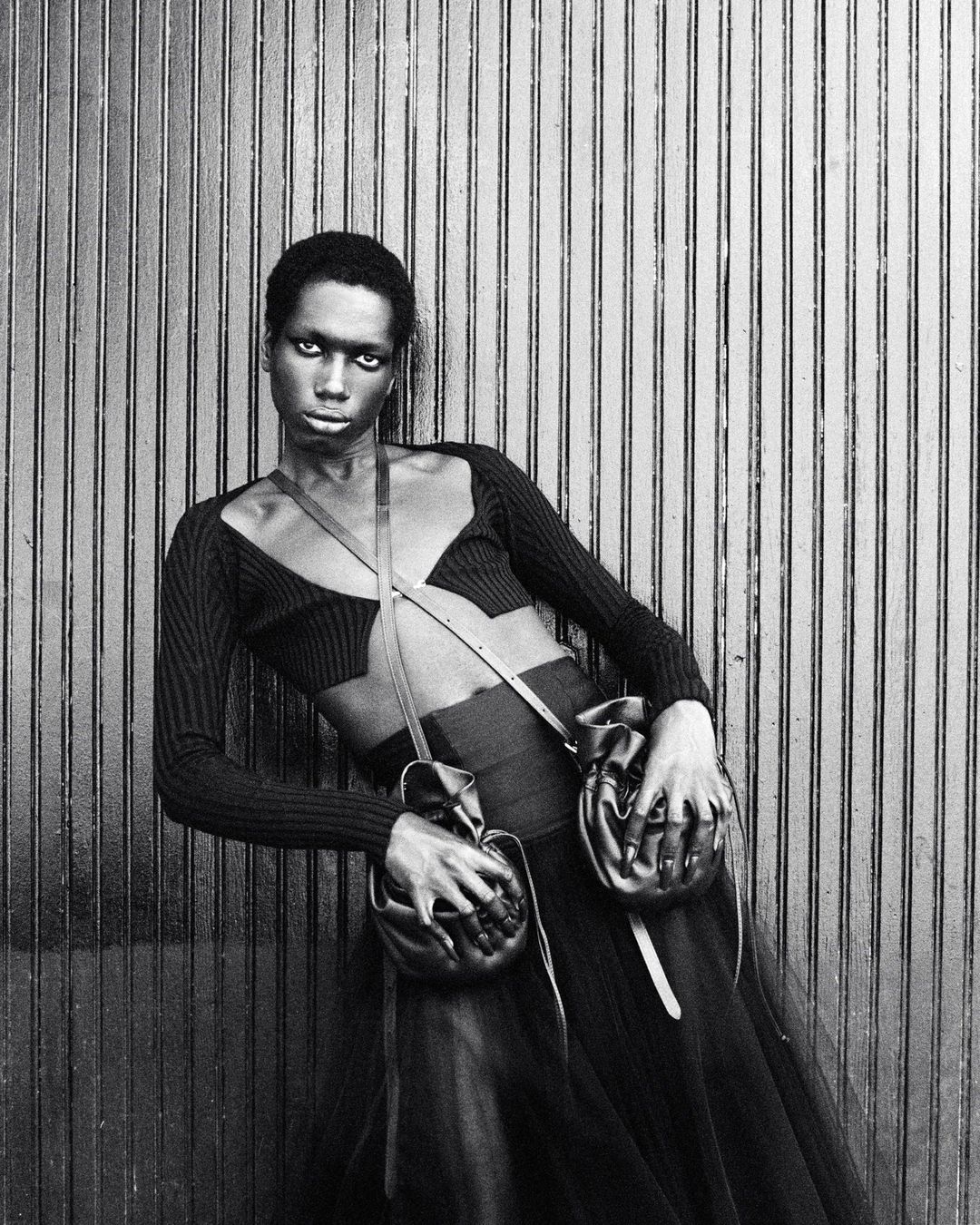 A Black fashion model poses leaning against a wall, wearing a black cut-out top, skirt, and two leather pouches attached to a thin strap.