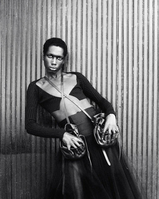 A Black fashion model poses leaning against a wall, wearing a black cut-out top, skirt, and two leather pouches attached to a thin strap.