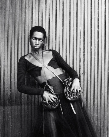 A Black fashion model poses leaning against a wall, wearing a black cut-out top, skirt, and two leather pouches attached to a thin strap.