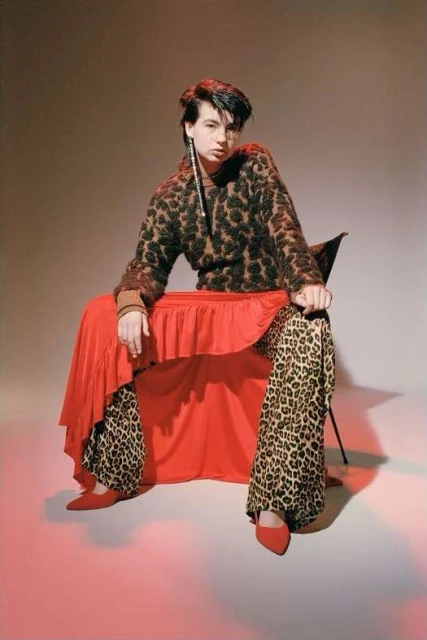 Adult female fashion model poses sitting on a black chair in a photo studio wearing leopard print sweater, pants, a red skirt and matching shoes.