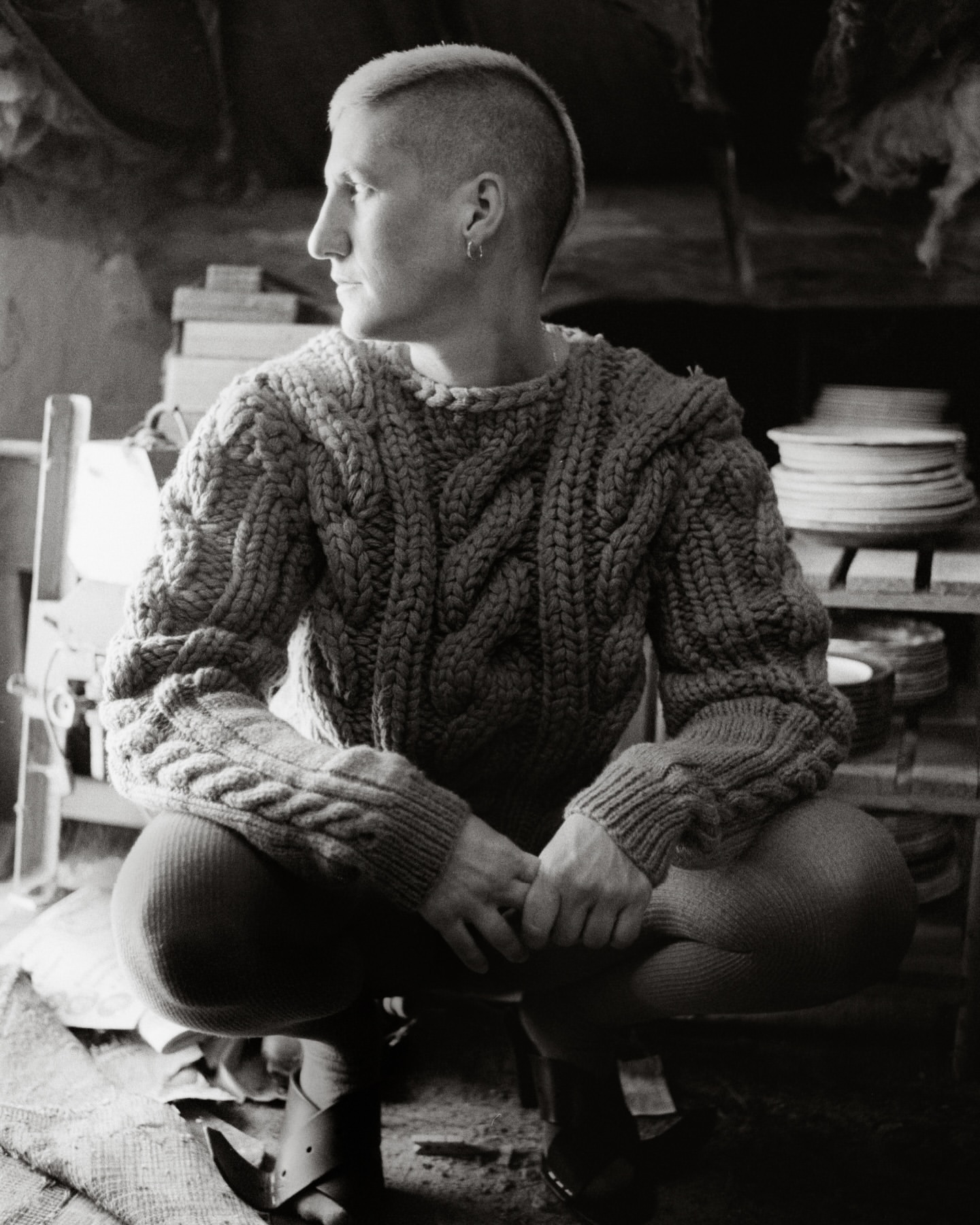 Black and white portrait of a person with a short mohawk haircut wearing a knit sweater, crouching indoors, looking to the side.