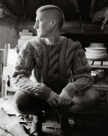 Black and white portrait of a person with a short mohawk haircut wearing a knit sweater, crouching indoors, looking to the side.