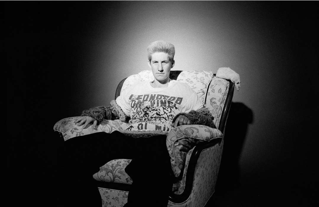 Black and white portrait of a man with a retro hairstyle wearing a graphic T-shirt and sitting in a floral patterned armchair.