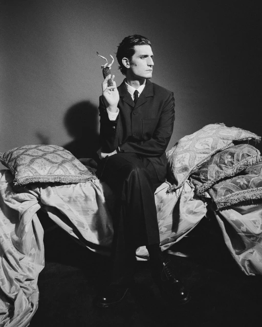 Black and white studio portrait of a male in a suit sitting on a bed with decorative pillows and holding a small figurine.