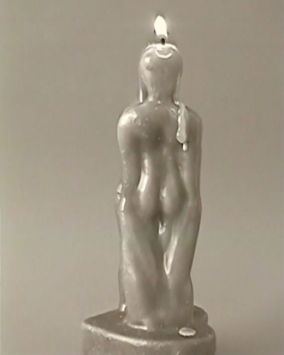 Burning figure candle with a wax buildup on its back stands against a neutral gray backdrop, its flame flickering on top.