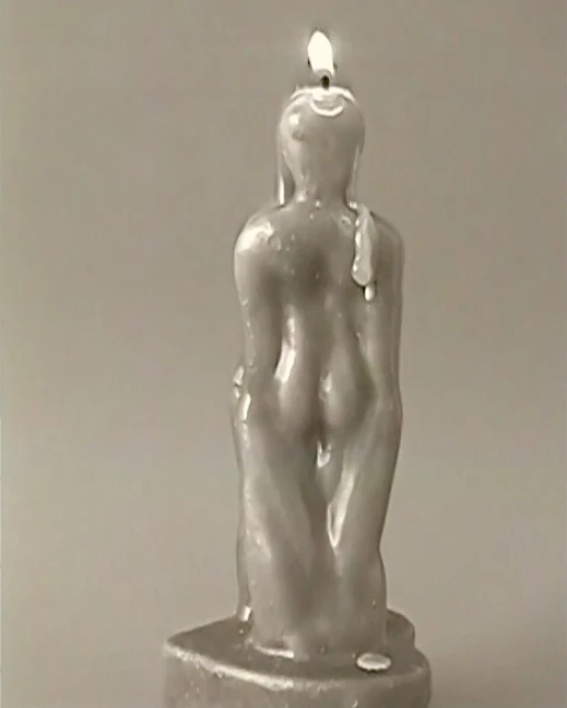 Burning figure candle with a wax buildup on its back stands against a neutral gray backdrop, its flame flickering on top.