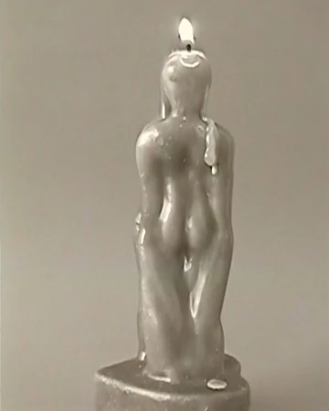 Burning figure candle with a wax buildup on its back stands against a neutral gray backdrop, its flame flickering on top.