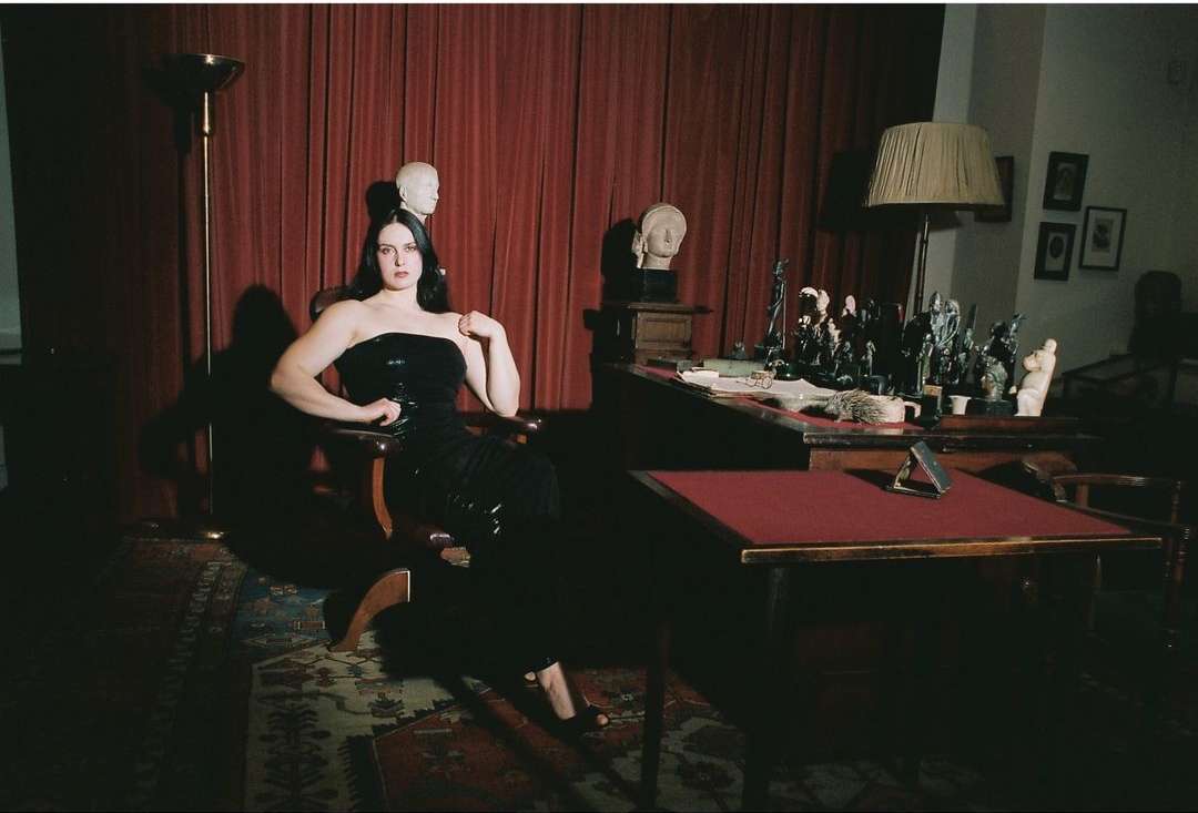 Woman in a black dress poses in a chair next to a desk with sculptures and busts. The setting is in a dimly lit room.