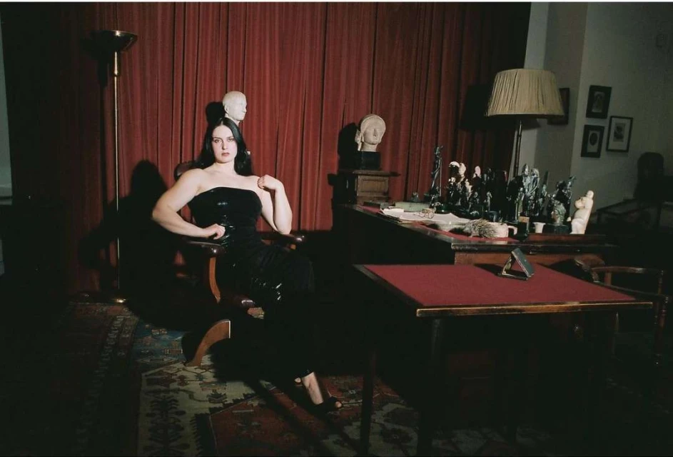 Woman in a black dress poses in a chair next to a desk with sculptures and busts. The setting is in a dimly lit room.