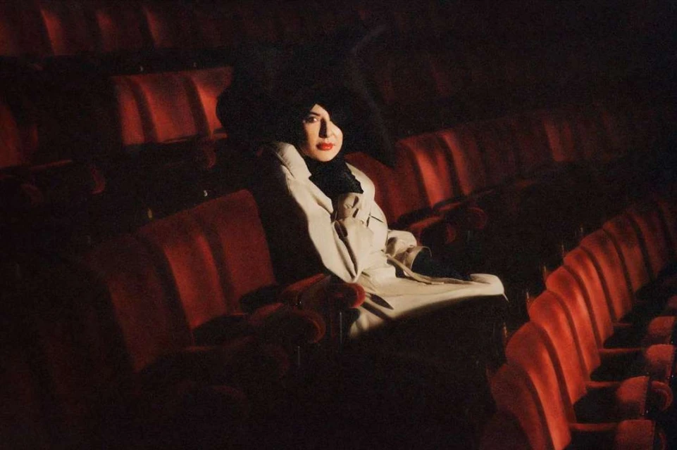 A woman in a cream-colored coat and black hat sits in a dimly lit theater with red seats, creating a dramatic and elegant atmosphere.