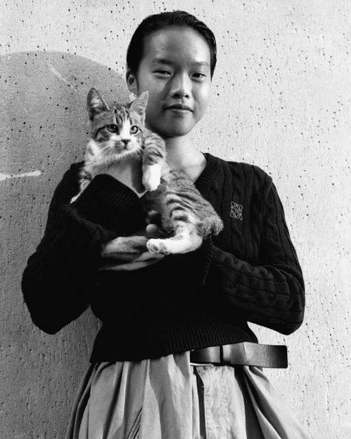 Monochrome portrait of a person holding a tabby kitten, wearing a v-neck sweater, wide skirt, and belt in front of a concrete wall.