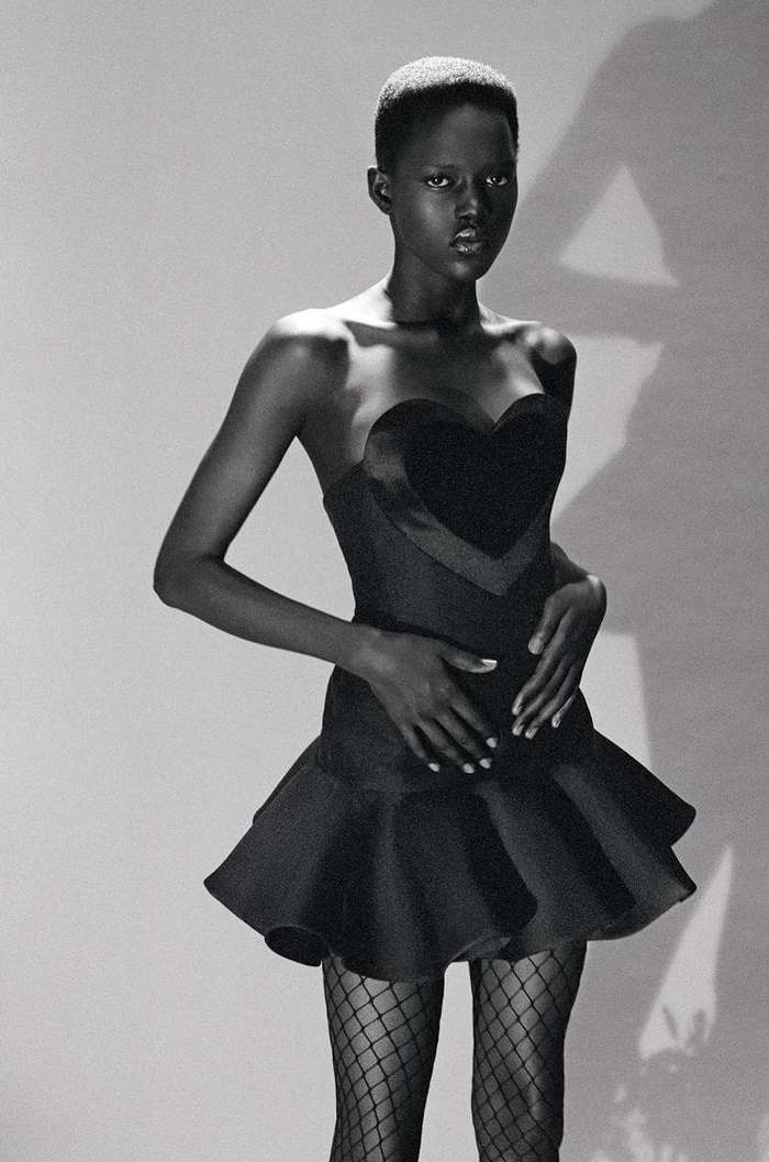 Black and white studio shot of a Black female model in a strapless heart-shaped dress and fishnet stockings posing against a plain backdrop.