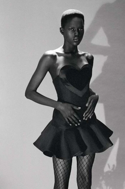 Black and white studio shot of a Black female model in a strapless heart-shaped dress and fishnet stockings posing against a plain backdrop.