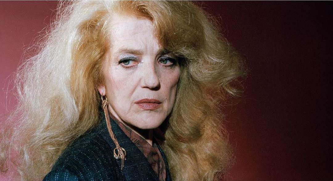 Close-up portrait of a mature fair-skinned woman with blonde hair, wearing long gold earrings against a dark reddish background.