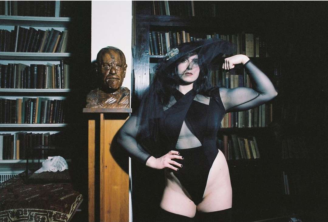 Woman poses indoors wearing black bodysuit and hat with veil, showcasing biceps, with books and sculpture in the background.