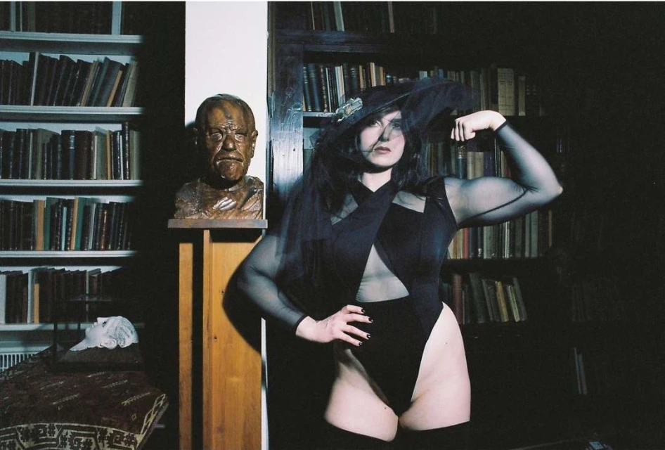 Woman poses indoors wearing black bodysuit and hat with veil, showcasing biceps, with books and sculpture in the background.